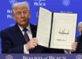 Trump holds an open signed document in front of a blue backdrop reading “Board of Peace.”